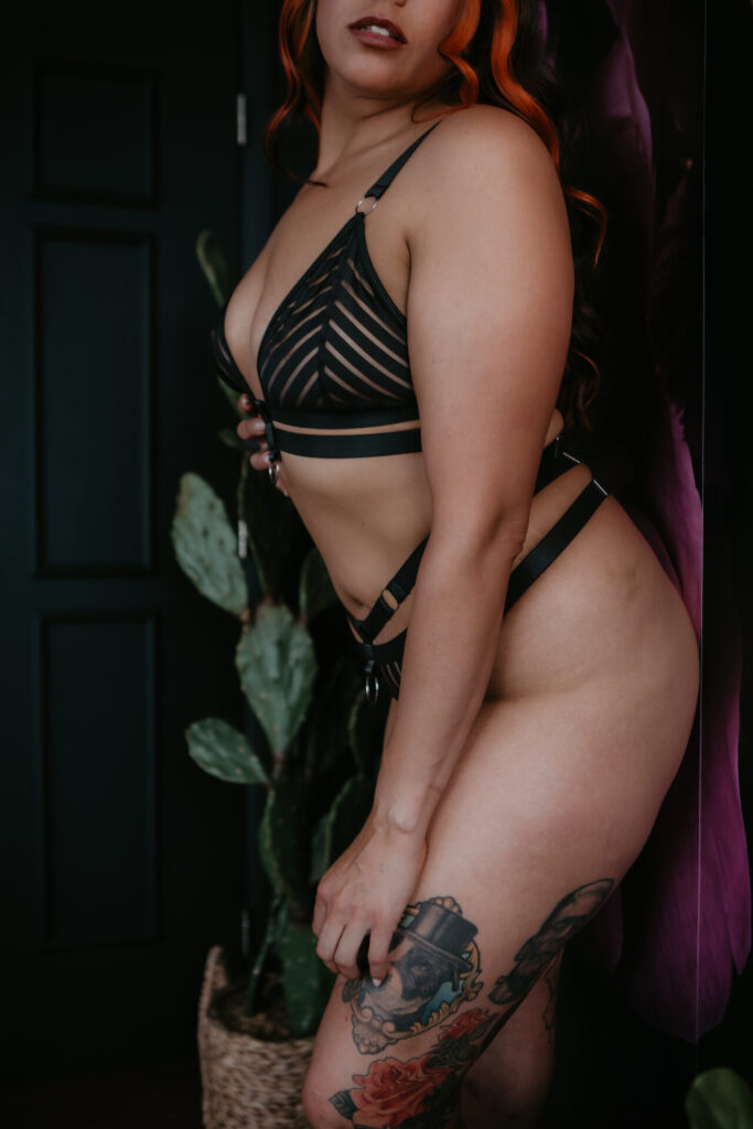Boudoir photo of a tattooed woman in black strappy lingerie posed by a cactus and dark floral backdrop at Arielle Roth’s Bay City studio.