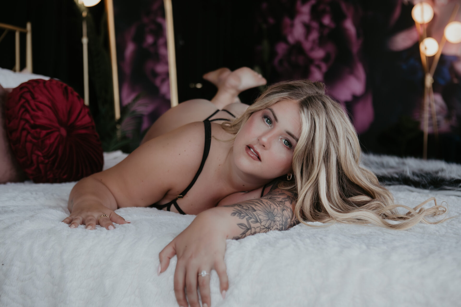 Boudoir photo of a blonde woman with tattoos lying on a white bed in black lingerie at Arielle Roth’s Bay City boudoir studio.