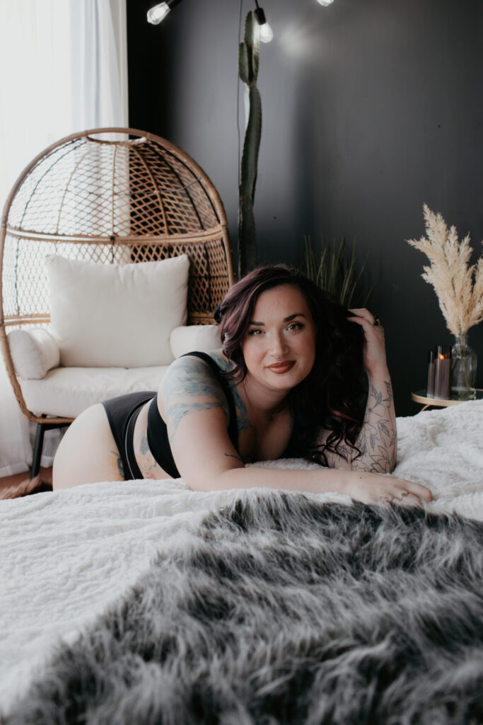 Boudoir photo of a confident woman lying on a bed in a modern, softly lit studio. She has tattoos on her arms, wavy dark hair, and is smiling warmly at the camera, creating an intimate and empowering vibe.