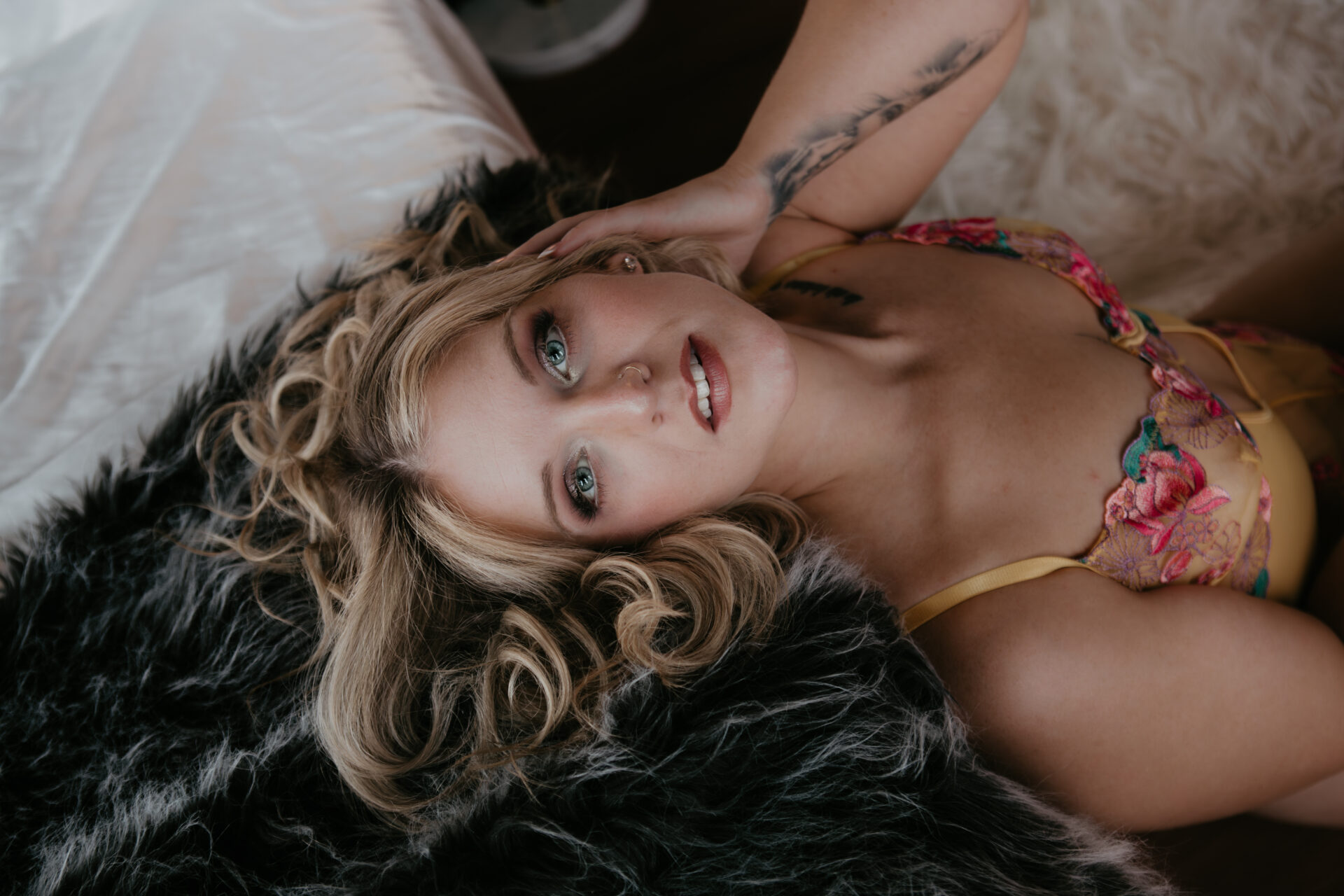Blonde woman with curled hair and soft glam makeup lying on a faux fur throw, wearing floral yellow lingerie, looking confidently into the camera.