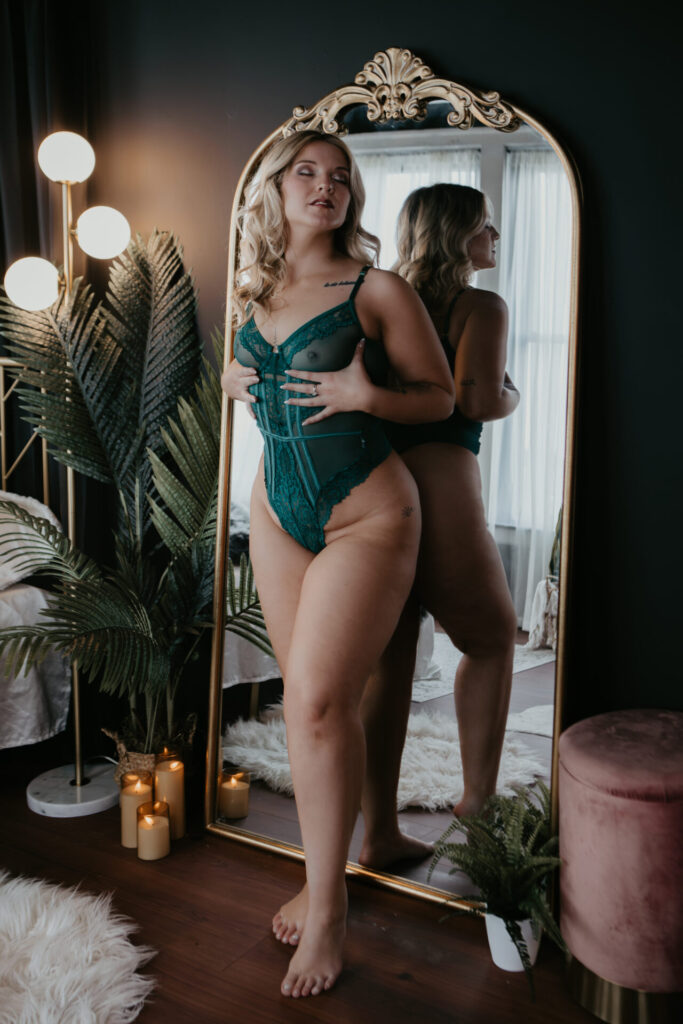 Woman in teal lingerie standing in front of a gold mirror with candles and plants during a boudoir session at Arielle Roth Boudoir in Bay City.