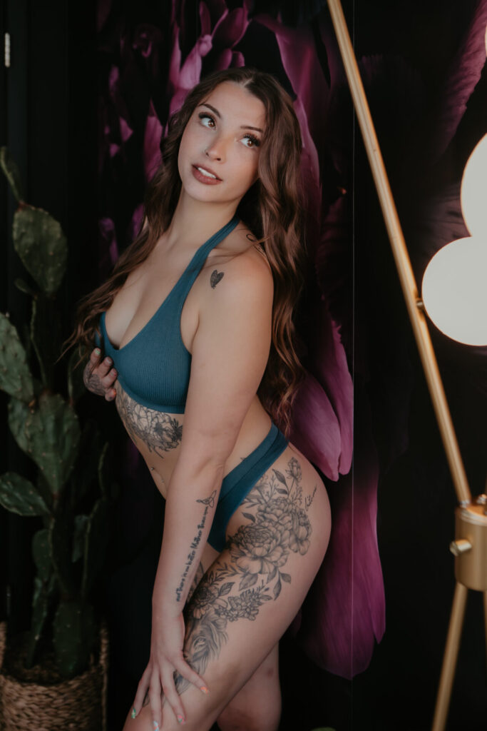 Confident woman in teal lingerie showing floral and lion tattoos during boudoir photoshoot in front of floral backdrop.