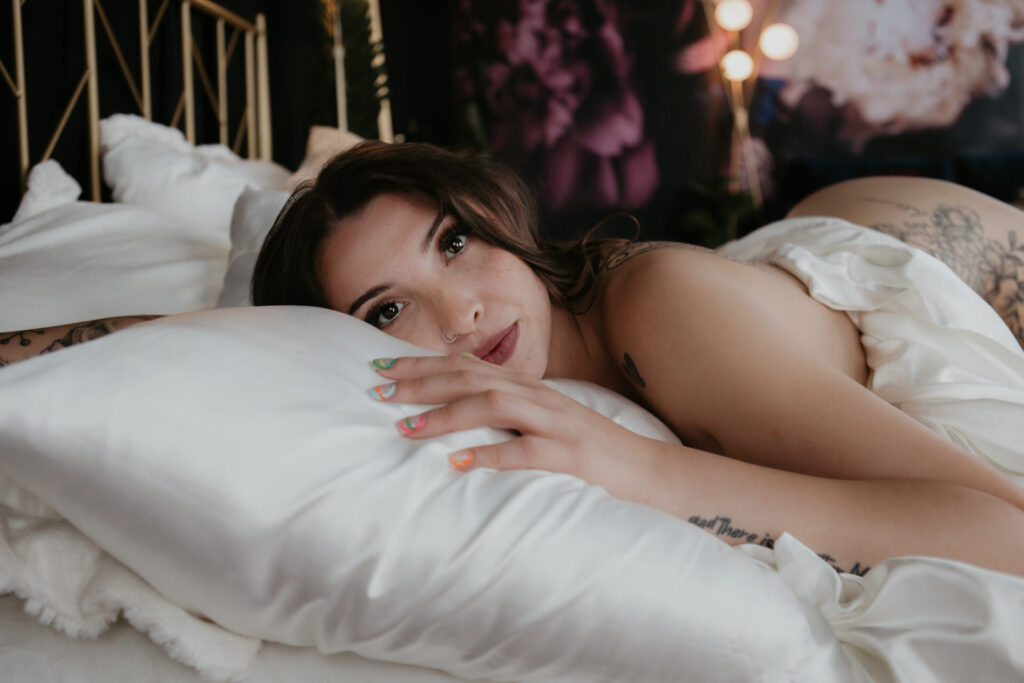 Tattooed woman laying on bed with silky sheets, gazing into the camera with colorful nails.