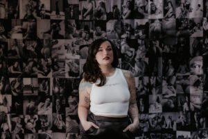Tattooed boudoir photographer in white crop top standing confidently in front of a collage of sensual black and white boudoir portraits