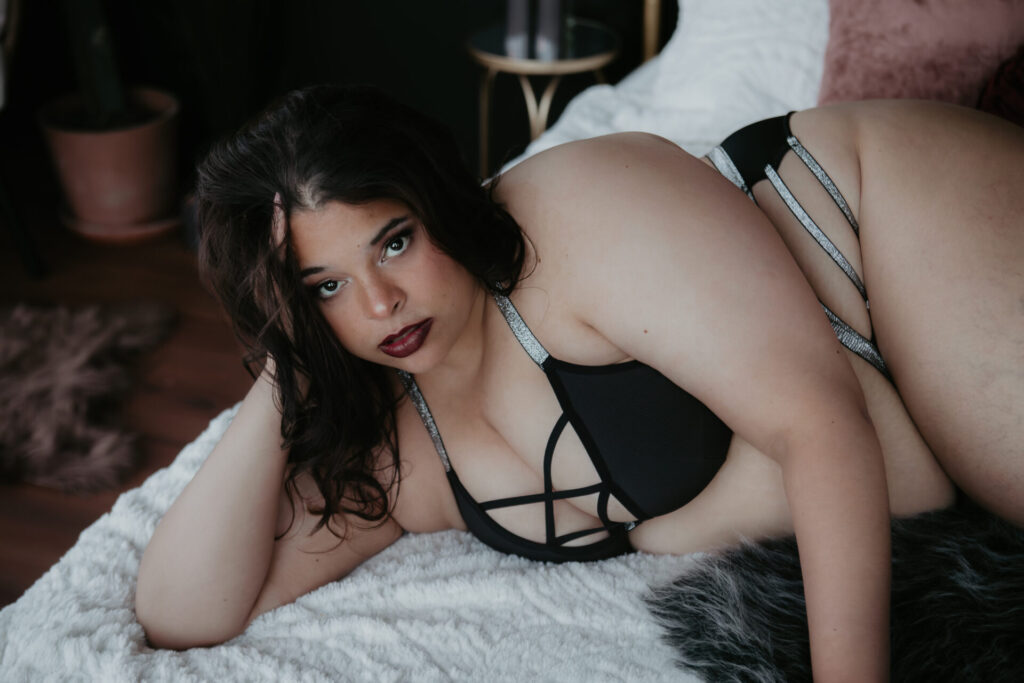 Plus-size woman in strappy black lingerie lying on a bed, gazing confidently into the camera during a boudoir session.