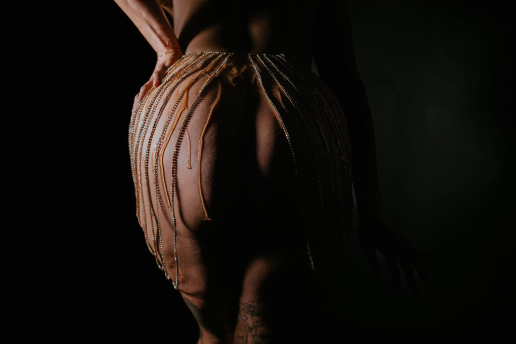 Back view of a woman's body with honey and crystal chains dripping over her curves during a boudoir session.