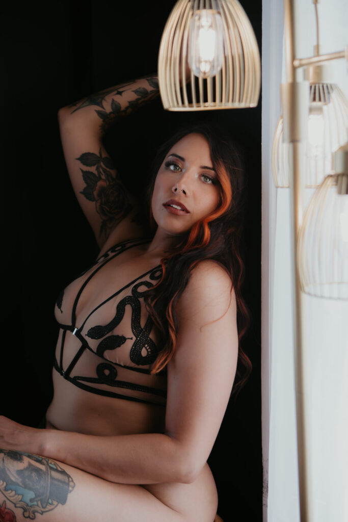 Tattooed woman with long brown and orange-highlighted hair posing in black sheer lingerie with snake detailing, seated confidently by a window with modern pendant lights in a luxury boudoir studio.