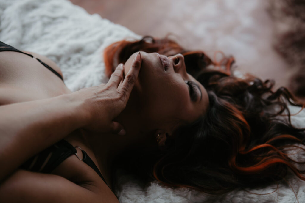 Woman with long wavy hair and orange highlights lying on her back in black lingerie, softly touching her jawline with manicured fingers in a sensual boudoir pose on a white blanket.