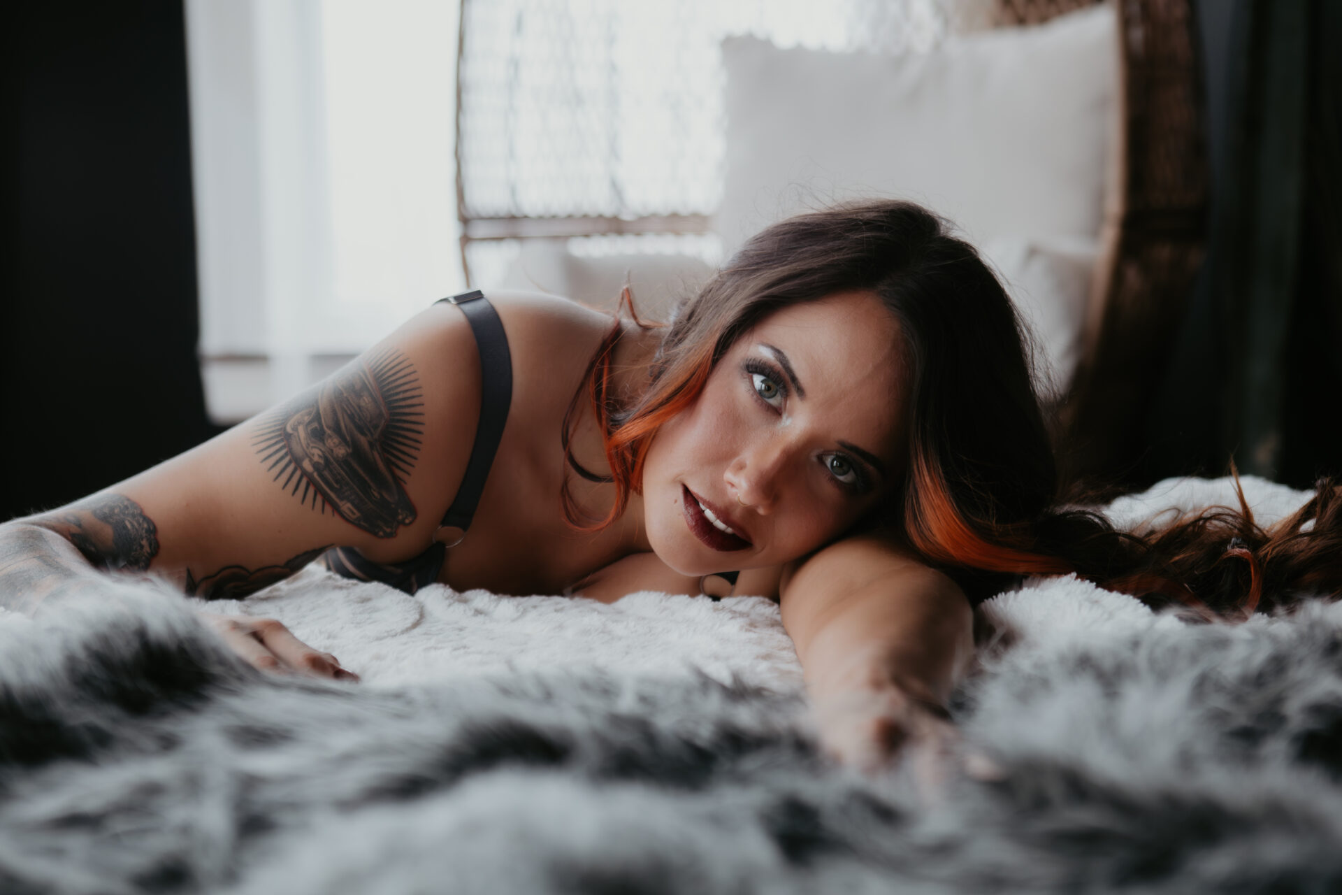 Tattooed woman with long brunette and copper-highlighted hair lying on a white fur bed, gazing confidently into the camera in a private boudoir studio setting.