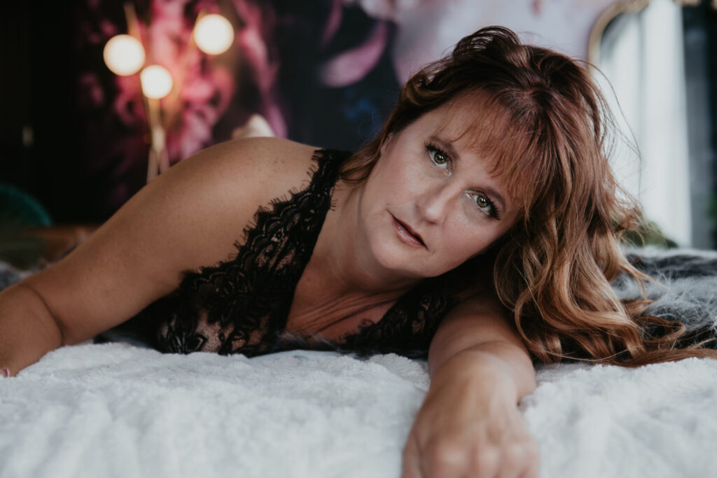 Mature boudoir client over 50 from Alpena, Michigan lying on the bed in black lace lingerie during her empowering session at Arielle Roth Boudoir studio in Bay City.