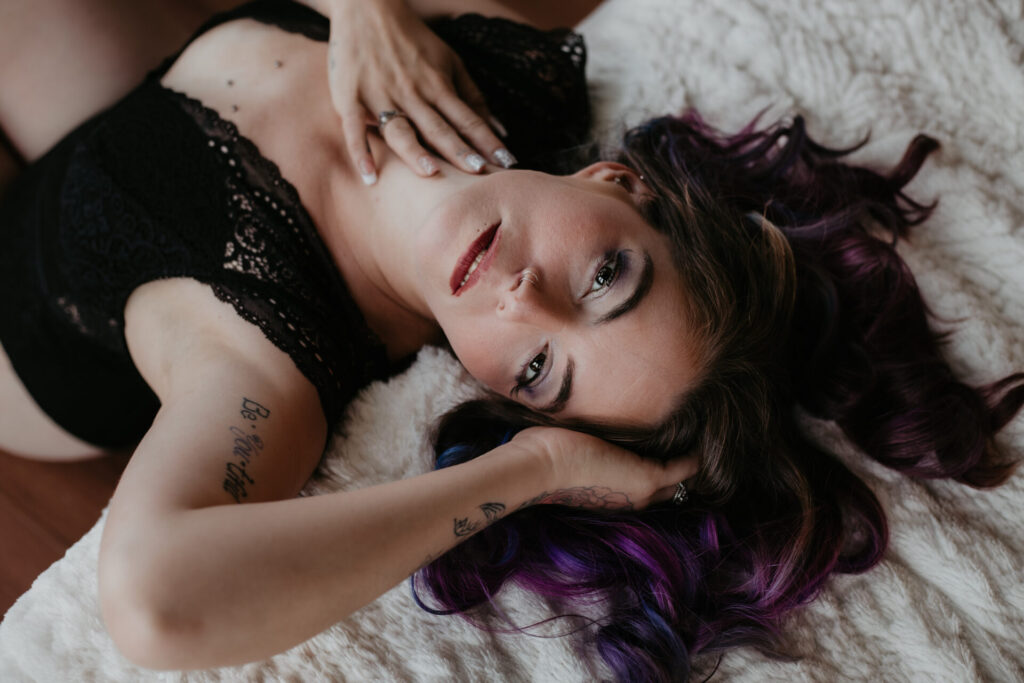Michigan boudoir photography session with woman in black lace lingerie, lying back with purple hair and confident gaze.