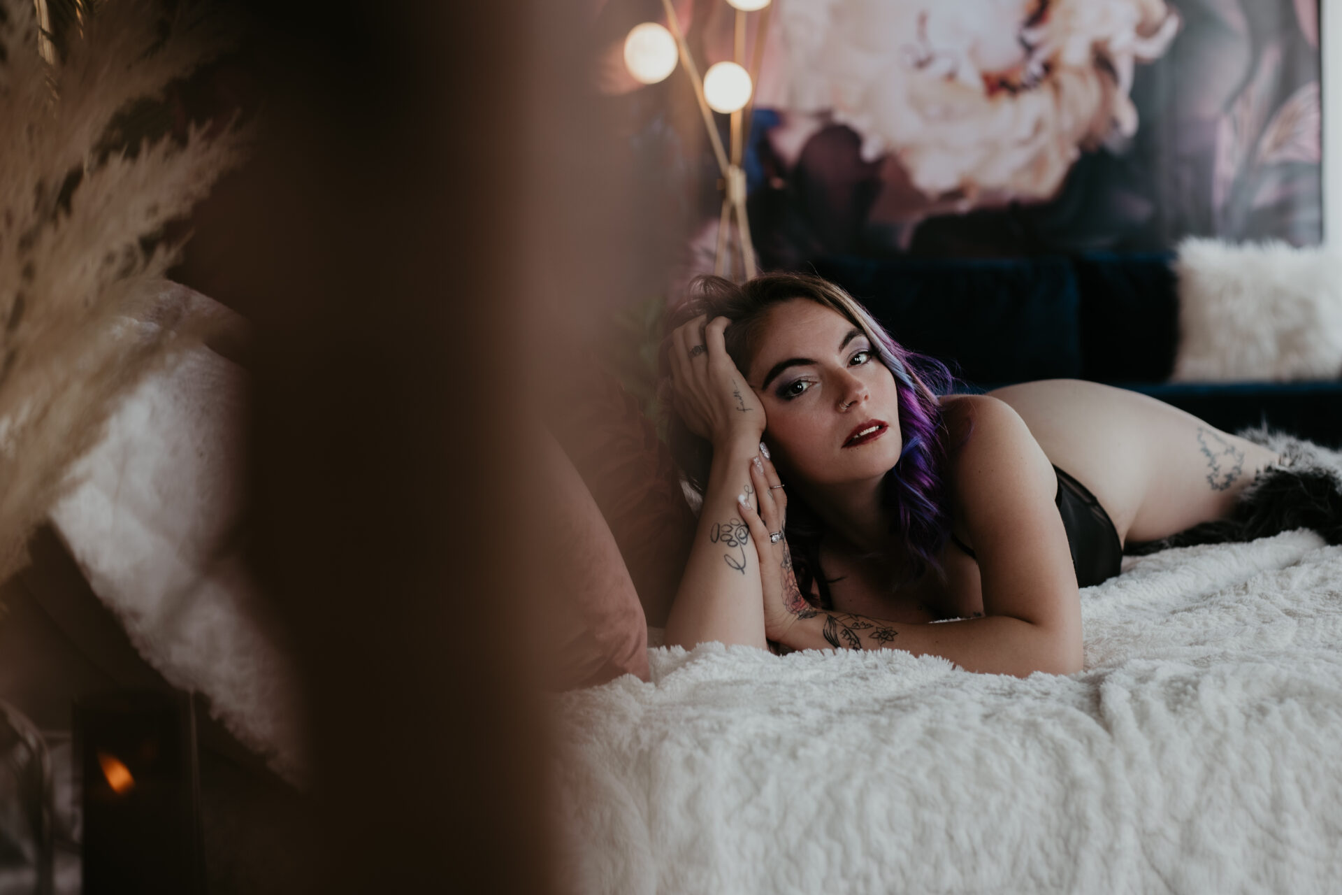 Michigan boudoir photography of woman with purple hair in black lingerie, lying on bed with soft gaze and tattoos visible.