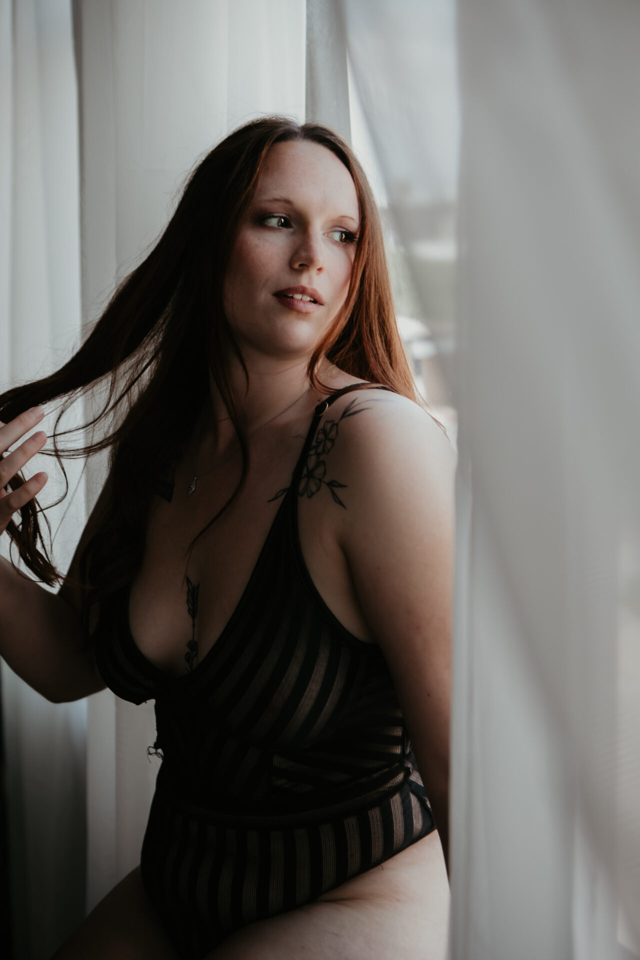 Boudoir portrait of a woman in black striped lingerie softly lit by natural window light, playing with her hair and gazing to the side.