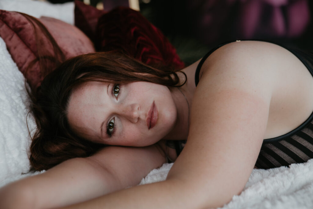 Close-up boudoir portrait of client from West Branch, Michigan with soft makeup and natural red hair, laying on white bedding in Bay City studio.