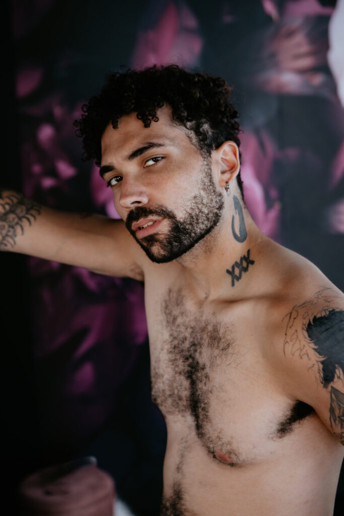 Shirtless male boudoir client with tattoos posing confidently in front of floral backdrop