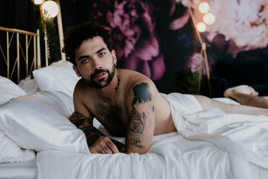 Detroit male boudoir client laying shirtless on bed with white sheets and tattoos on display