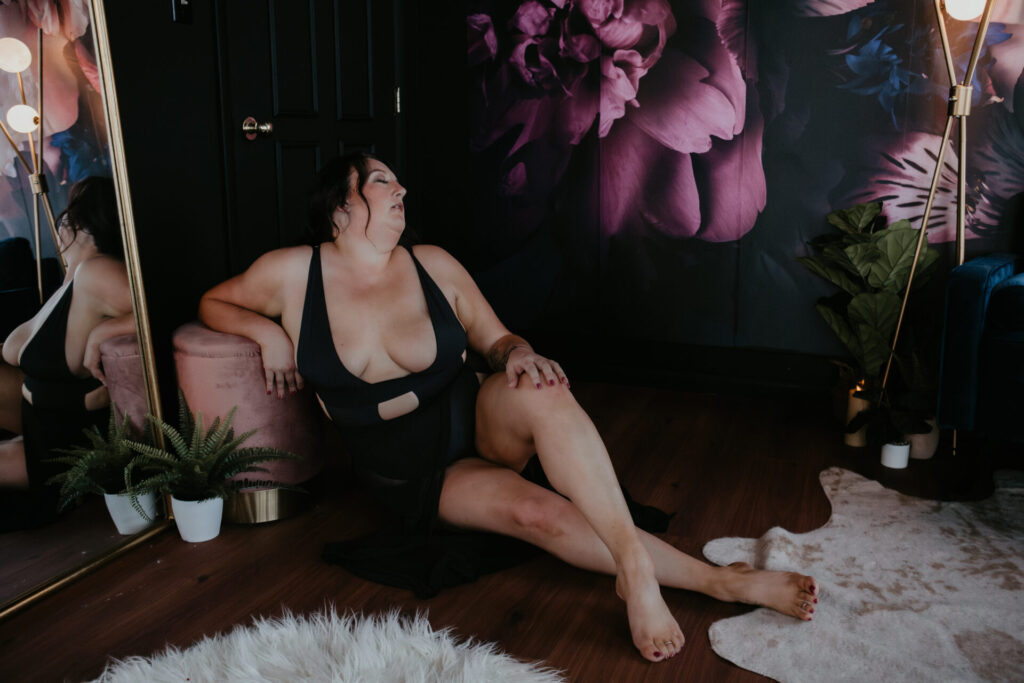 Plus-size woman in black lingerie lounging on the floor beside a mirror at Arielle Roth Boudoir studio in Bay City, Michigan.