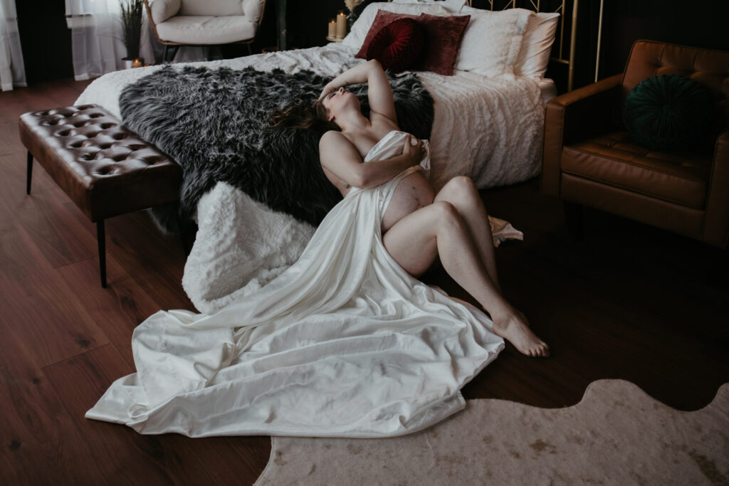 Maternity boudoir photo in Bay City, Michigan featuring a pregnant woman draped in white satin fabric, reclining on the floor near a bed.