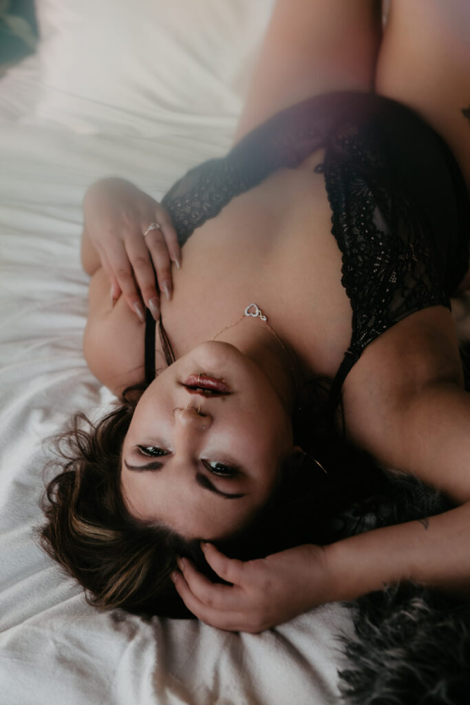 Boudoir photography Bay City Michigan woman in black lace lingerie lying on bed with heart necklace and confident gaze