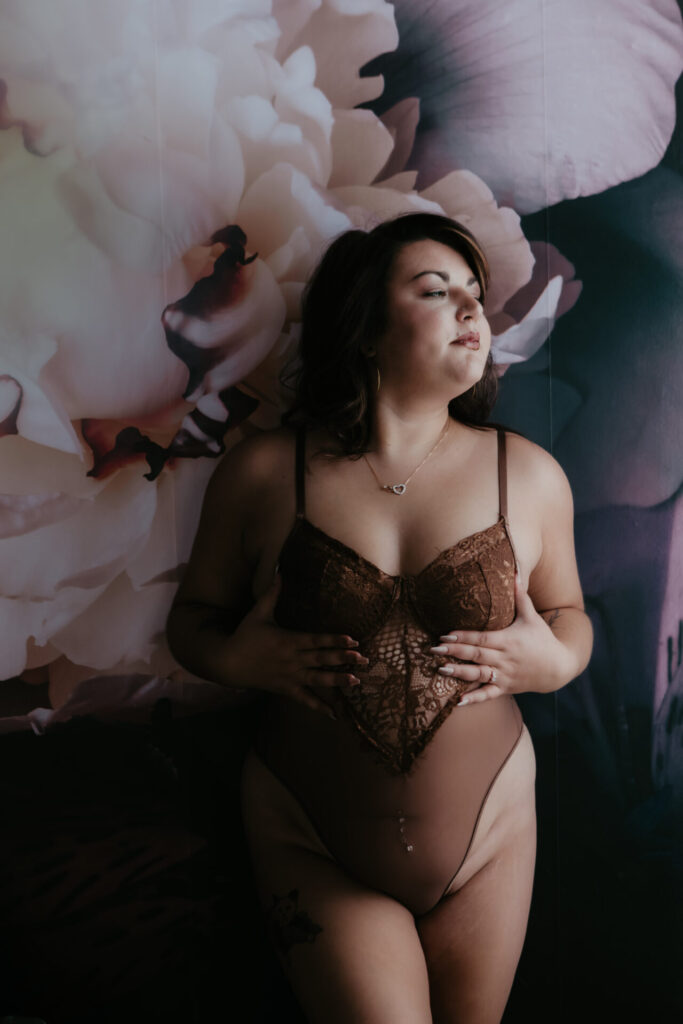 Boudoir photography Bay City Michigan woman in brown lace lingerie standing against floral wall with natural light