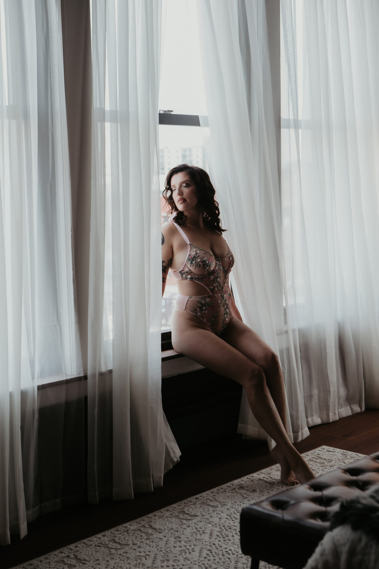 Boudoir photo of a Coleman, Michigan client in a pink floral bodysuit sitting on a window ledge with sheer curtains, photographed in my Bay City boudoir studio.