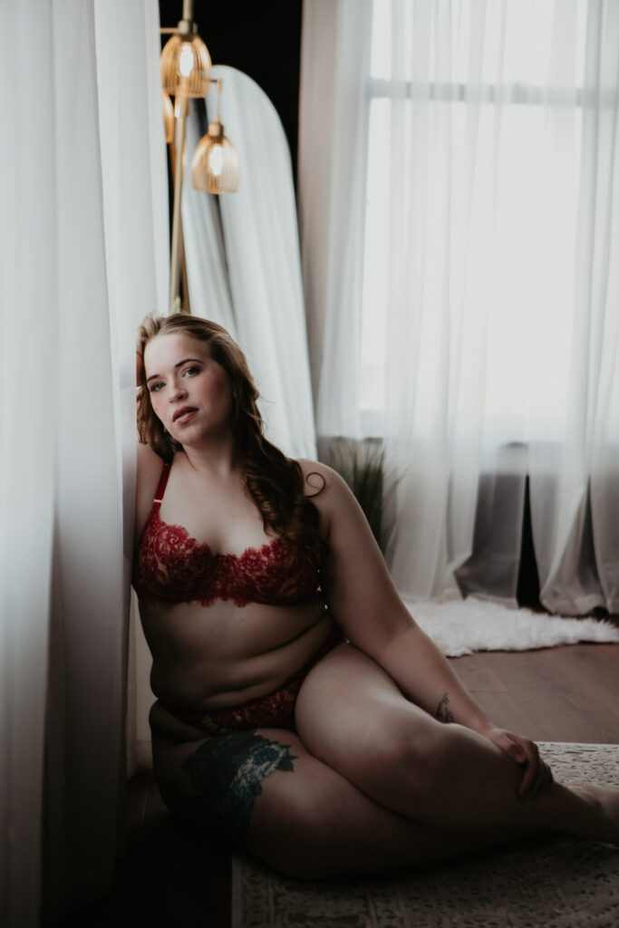 Boudoir portrait of a Saginaw client sitting by the window in red lace lingerie at Arielle Roth Boudoir studio in Bay City, Michigan