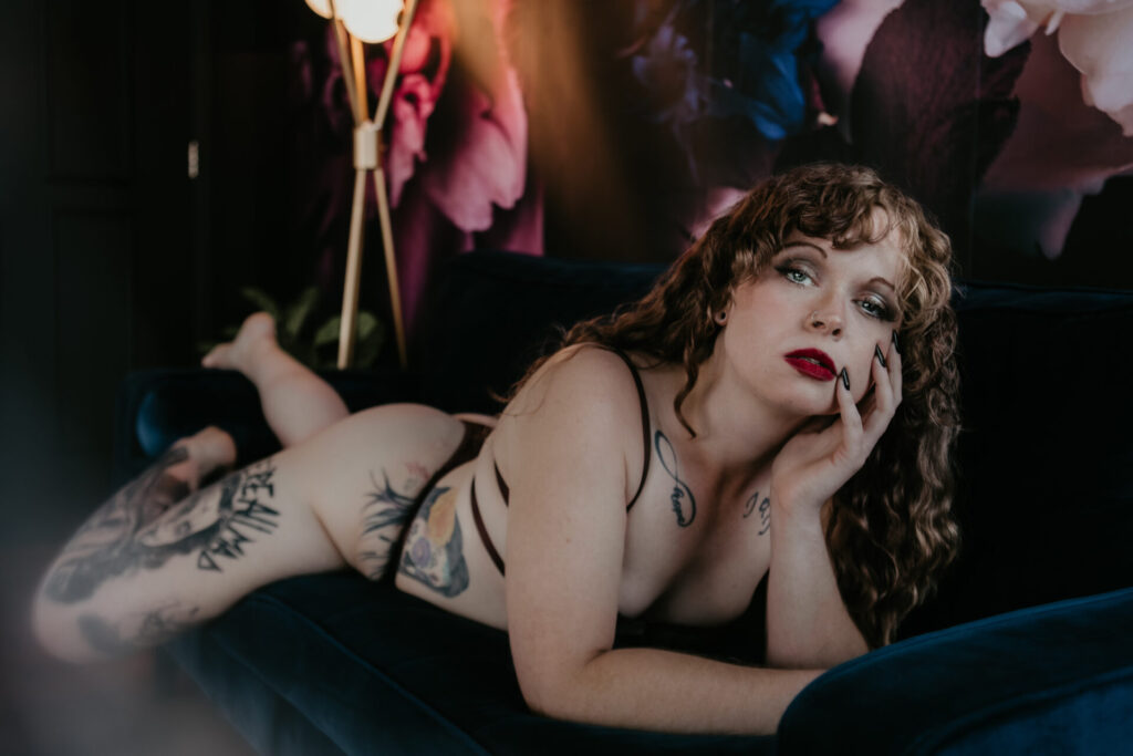Boudoir photo of a tattooed woman with red lips posing on a navy couch at Arielle Roth Boudoir studio in Bay City, Michigan