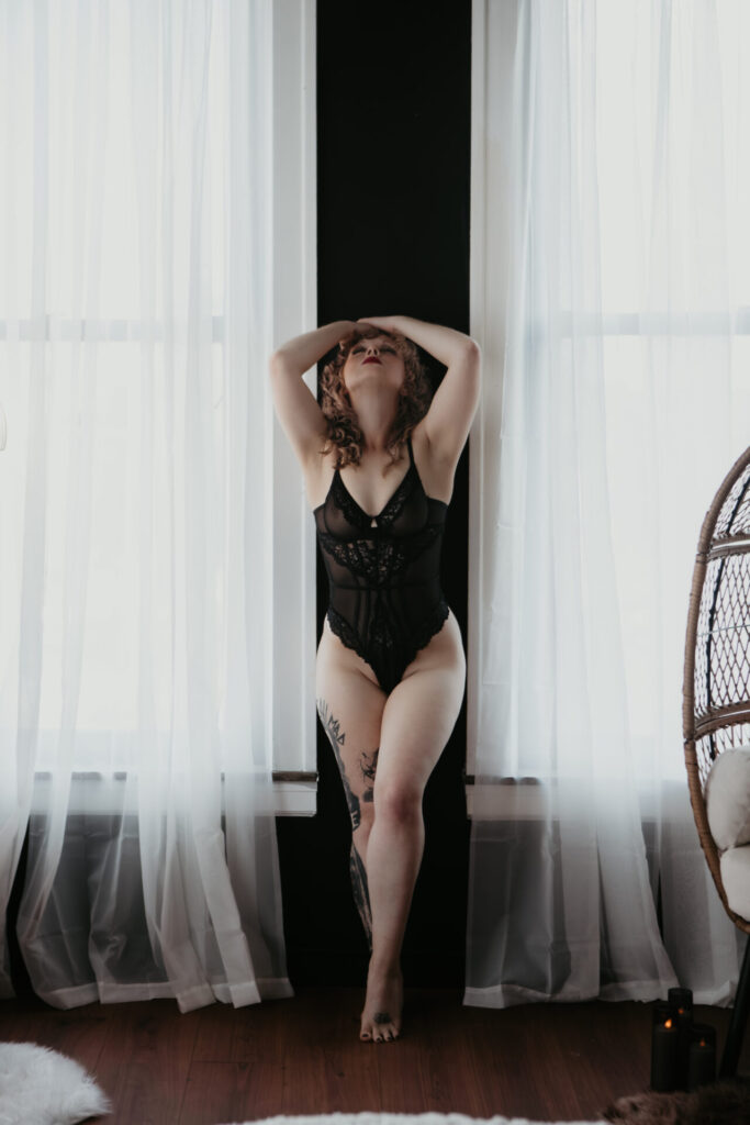 Boudoir portrait of a tattooed woman in black lace bodysuit posing between two windows with sheer curtains in Bay City Michigan studio