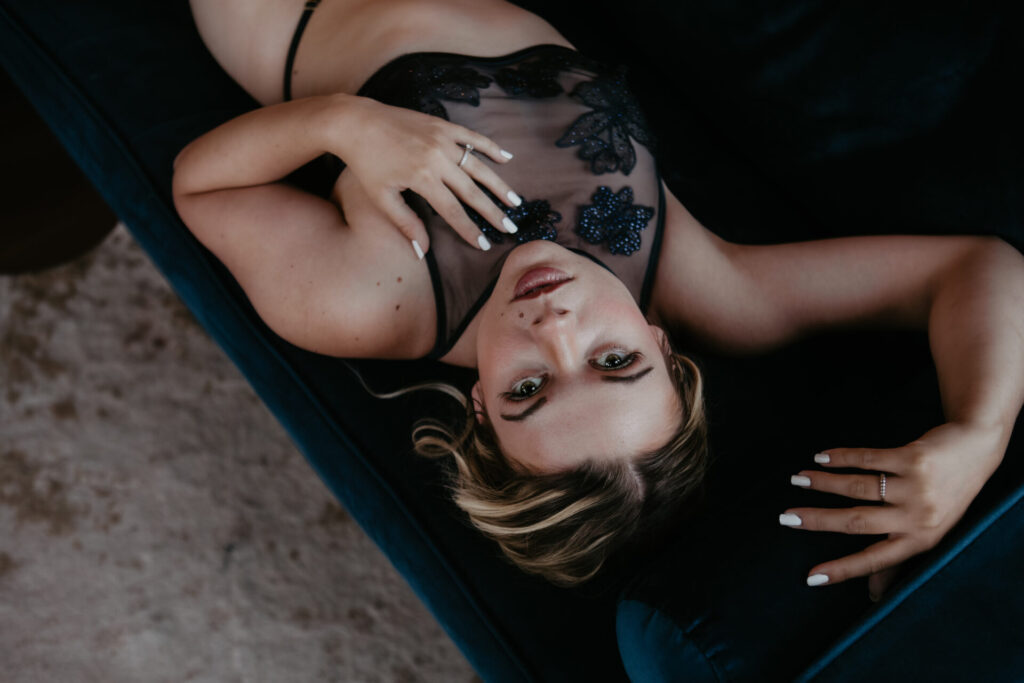 Boudoir portrait of a woman in sheer black floral lingerie reclining on a velvet couch during a Michigan boudoir session