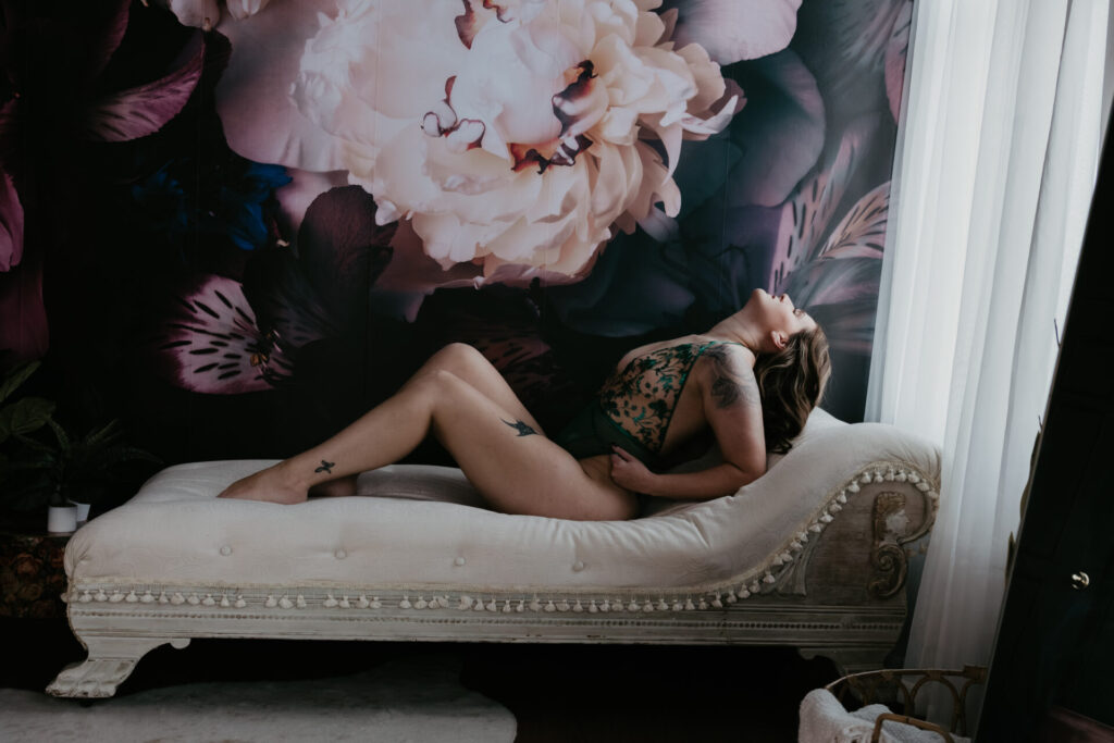 Woman in a green embroidered bodysuit arching back on a cream chaise lounge in a Bay City Michigan boudoir studio with a dark floral wall.