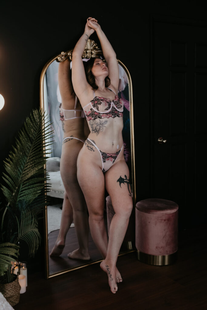 Woman standing on her toes in pink floral lingerie, arms stretched overhead, posing confidently in front of a tall gold mirror in a Bay City Michigan boudoir studio.