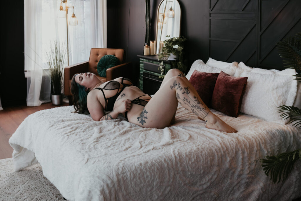 Woman in black fishnet lingerie lying on a white textured bed, showing tattoos and soft natural light inside a Bay City Michigan boudoir studio.