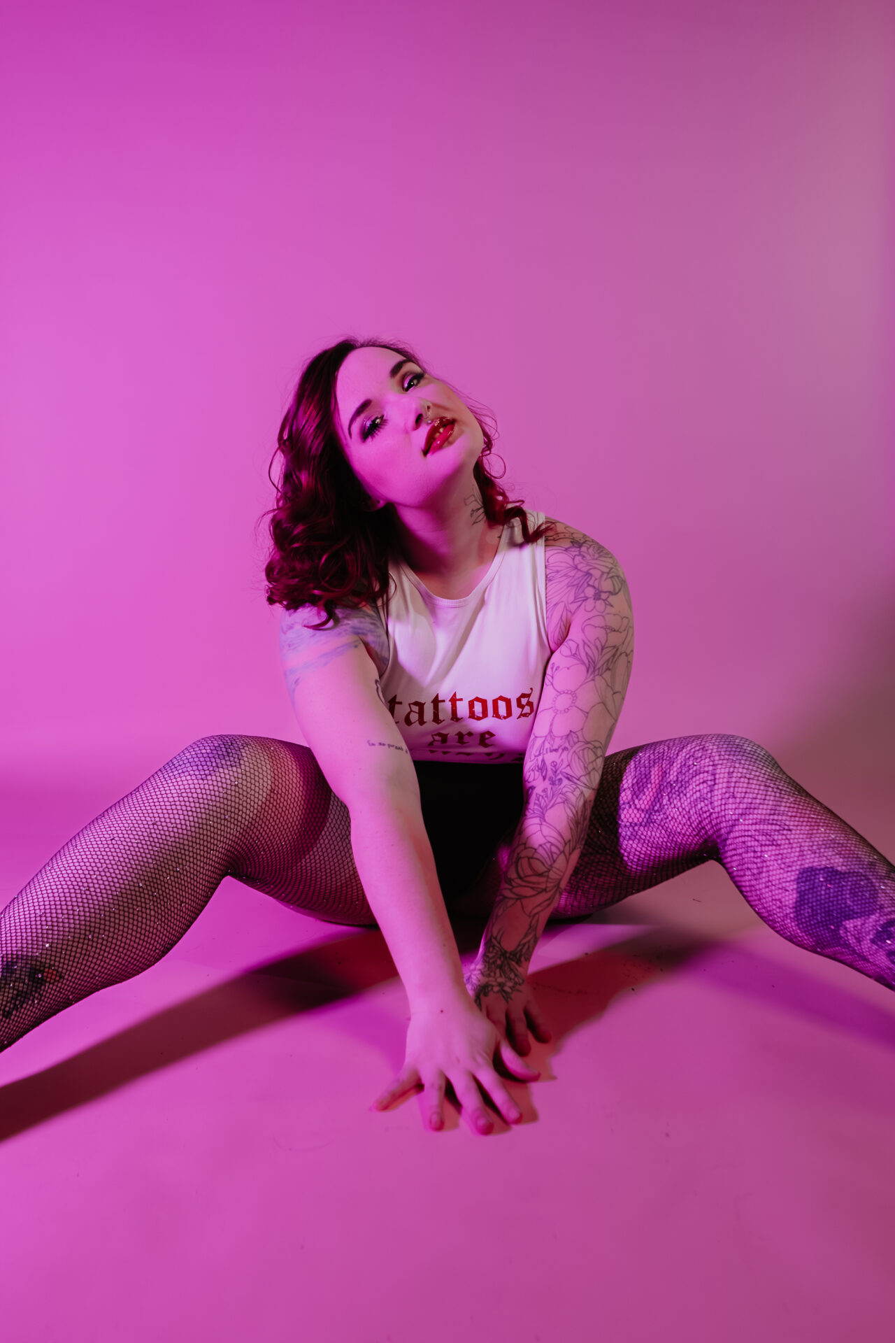 Tattooed boudoir client posed on the studio floor under pink lighting, wearing fishnet tights and a sleeveless top during a Bay City Michigan boudoir session.