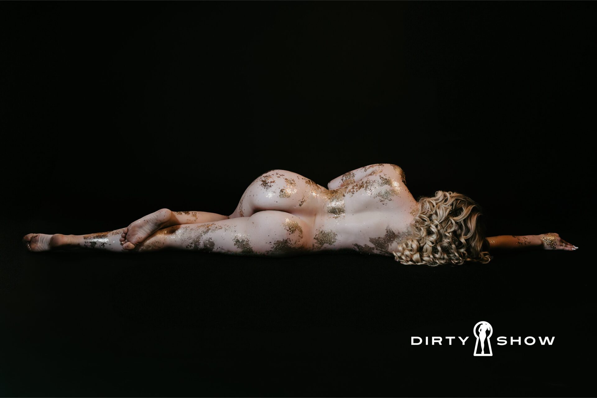 Fine art boudoir photograph of a nude figure reclined on her side with gold leaf accents on the body against a black background, featuring The Dirty Show logo, created by a Bay City, Michigan boudoir photographer.
