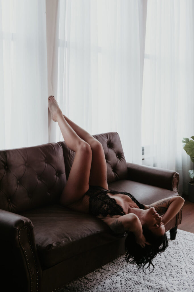 Boudoir photograph of a woman over 40 from Chesaning, Michigan reclining upside down on a leather couch in black lace lingerie, photographed in a Bay City Michigan boudoir studio.