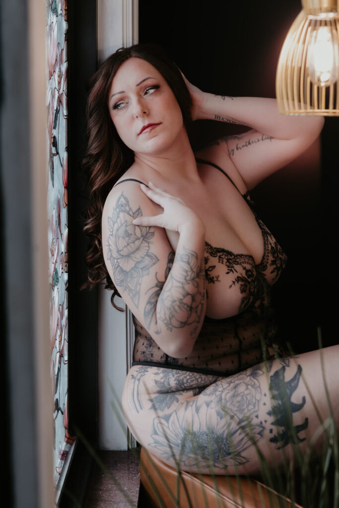 Woman seated by a window during a boudoir session in a Michigan studio, wearing black lace lingerie with visible tattoos, soft window light, and a calm reflective pose