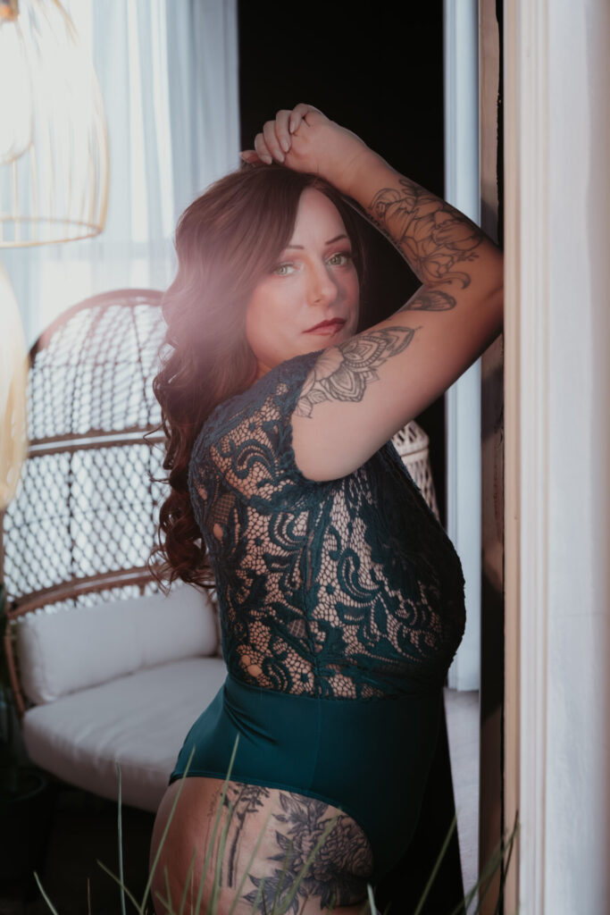 Color boudoir portrait of a woman posing near a doorway in a Michigan boudoir studio, teal lace bodysuit, arm raised above her head, tattoos visible, soft natural light