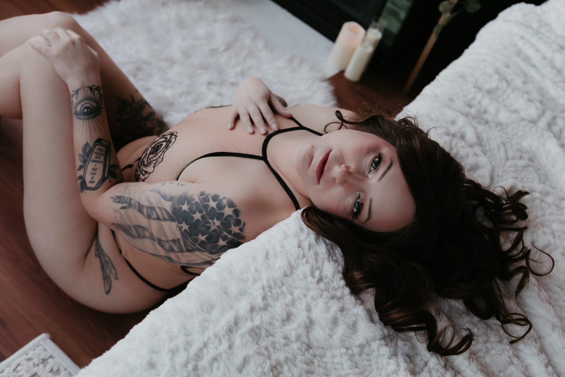 Color boudoir portrait of a tattooed woman lying on a white bed in a Michigan boudoir studio, looking directly at the camera, soft natural light highlighting her curves and relaxed expression