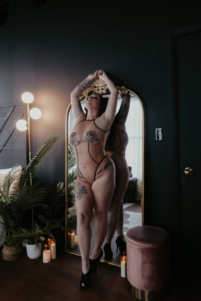 Full-body boudoir portrait of a tattooed woman standing in front of an ornate gold mirror with arms raised overhead, photographed in a Michigan boudoir studio with moody lighting and greenery