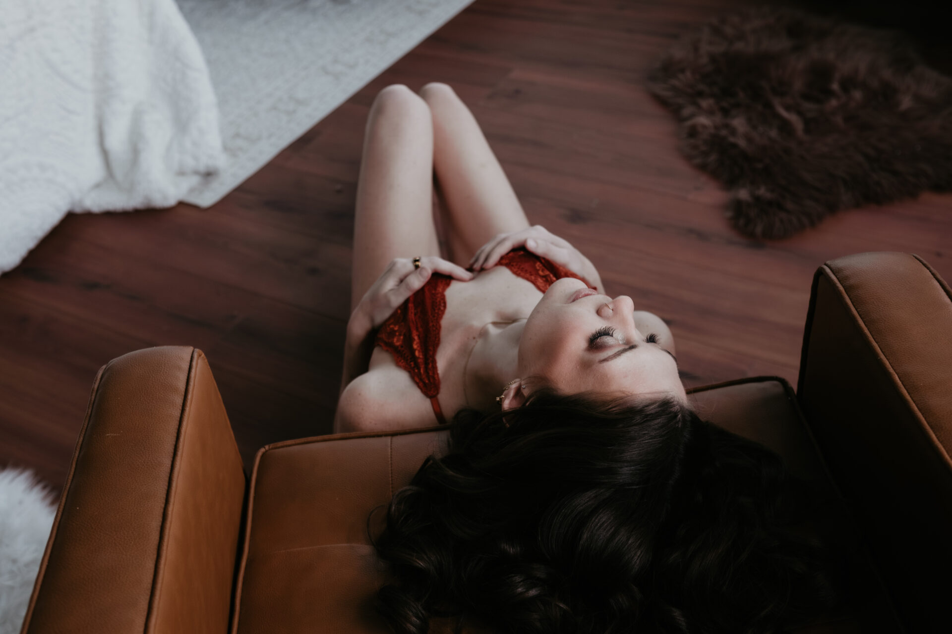 Boudoir portrait of a woman reclining on the floor between leather chairs in rust lace lingerie during a boudoir session in Bay City, Michigan. Client from Midland, Michigan.