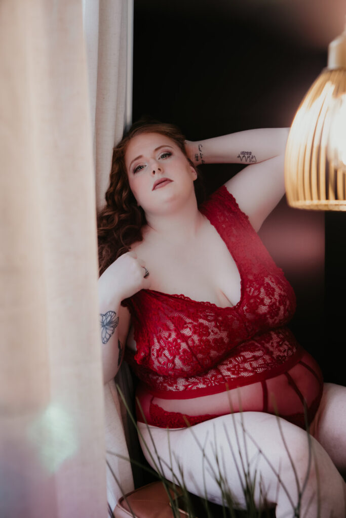 Plus size boudoir client from Vassar Michigan leaning against a window in red lace lingerie, photographed in soft studio light with an intimate indoor setting during a Michigan boudoir session.