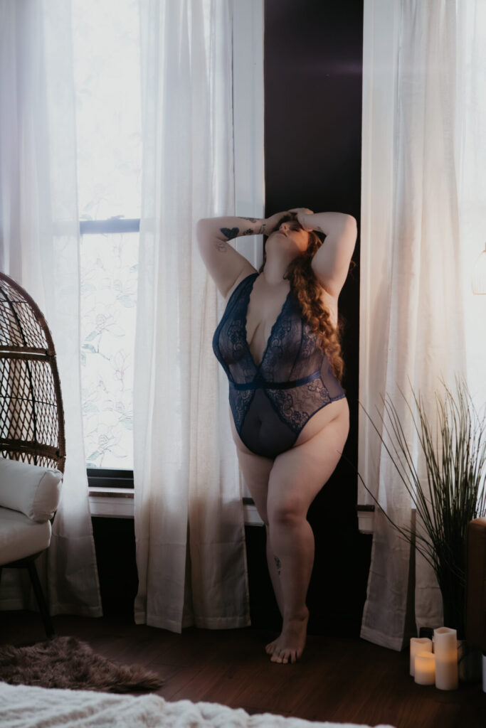Plus size boudoir client from Vassar Michigan standing between sheer curtains in blue lace lingerie, photographed in soft natural window light during a Michigan boudoir session.