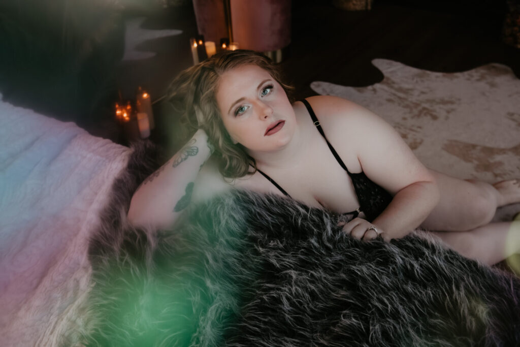 Color boudoir portrait of a plus size client reclining on a bed with faux fur textures, wearing black lingerie and looking directly at the camera in soft candlelight during a Michigan boudoir session.