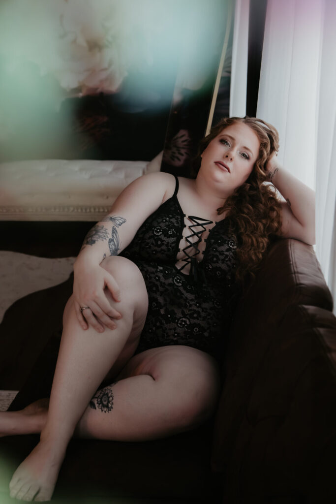 Color boudoir portrait of a plus size client seated on a leather couch near a window, wearing black lace lingerie and posing confidently during a Michigan boudoir photography session.