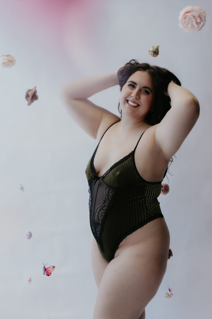 Boudoir portrait of a smiling client in a black lace bodysuit standing against a white backdrop with floating flowers and butterflies.