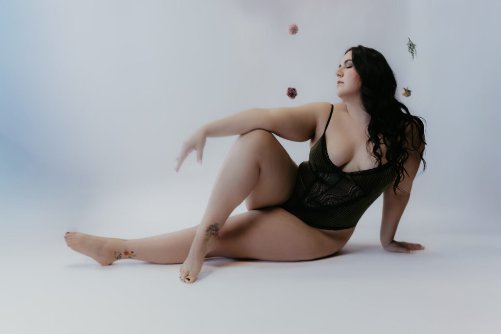 Boudoir portrait of a client in a black lace bodysuit seated on a white studio floor with one leg extended and floral accents in the background.