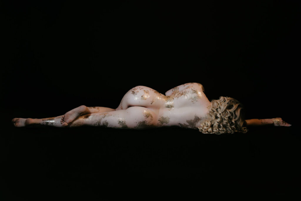 Fine art boudoir portrait of a nude woman lying on a black backdrop with gold leaf body art, photographed by Arielle Roth Boudoir in Michigan.