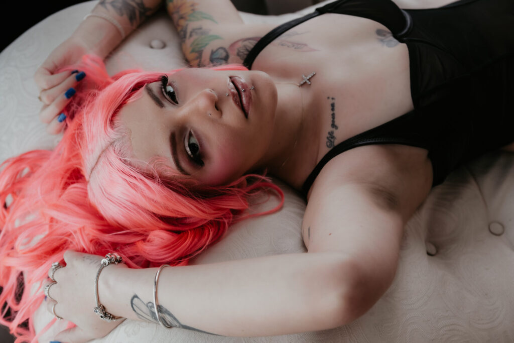 Woman with bright pink hair lying on a bed in black lingerie, showcasing tattoos and soft glam makeup during a boudoir session near Saginaw, Michigan