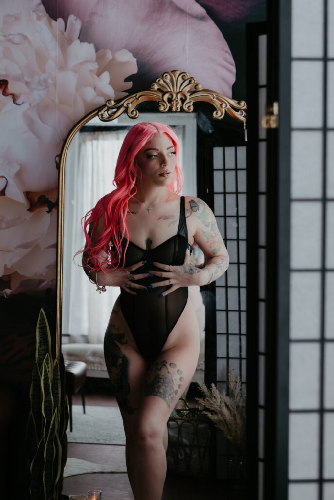 Woman with bright pink hair standing in front of a mirror in black lingerie during a boudoir session near Saginaw, Michigan
