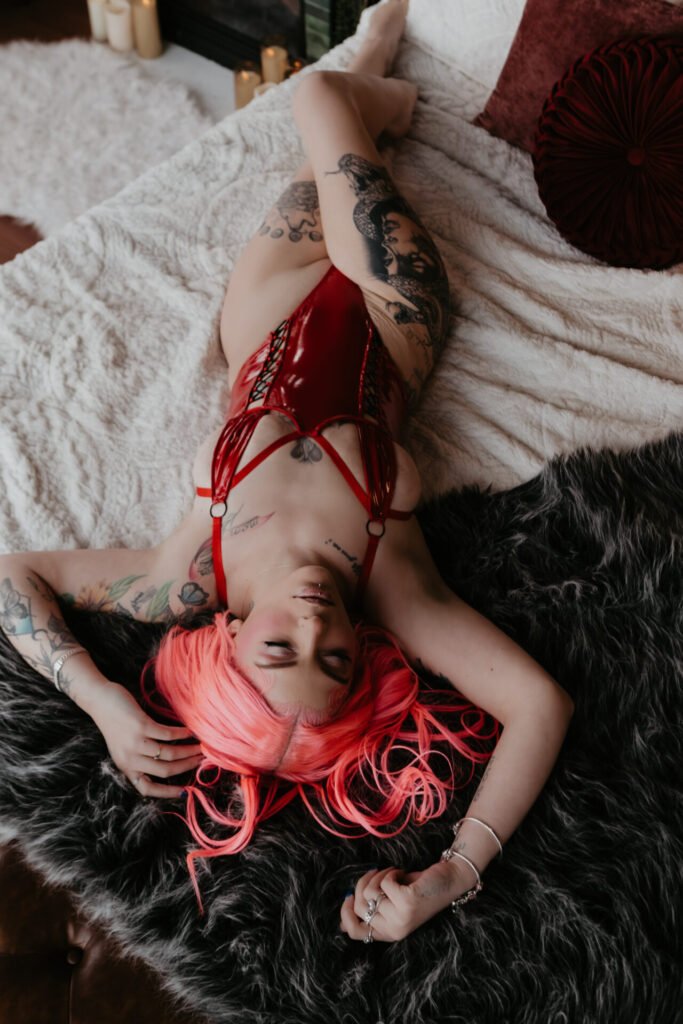 Overhead boudoir portrait of a woman with bright pink hair wearing red lingerie while lying on a bed during a session near Saginaw, Michigan